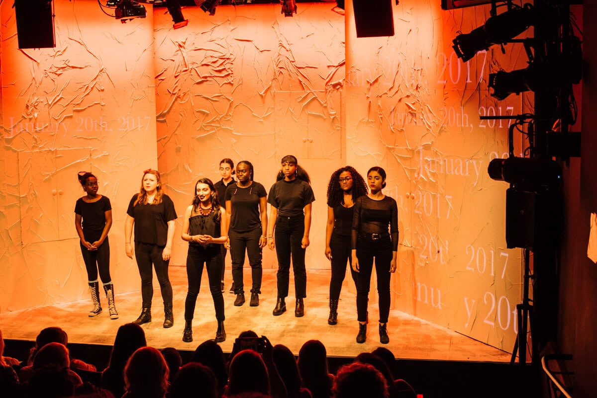 A group of performers stand onstage wearing all black. The stage is decorated in peeling paper and lit orange.